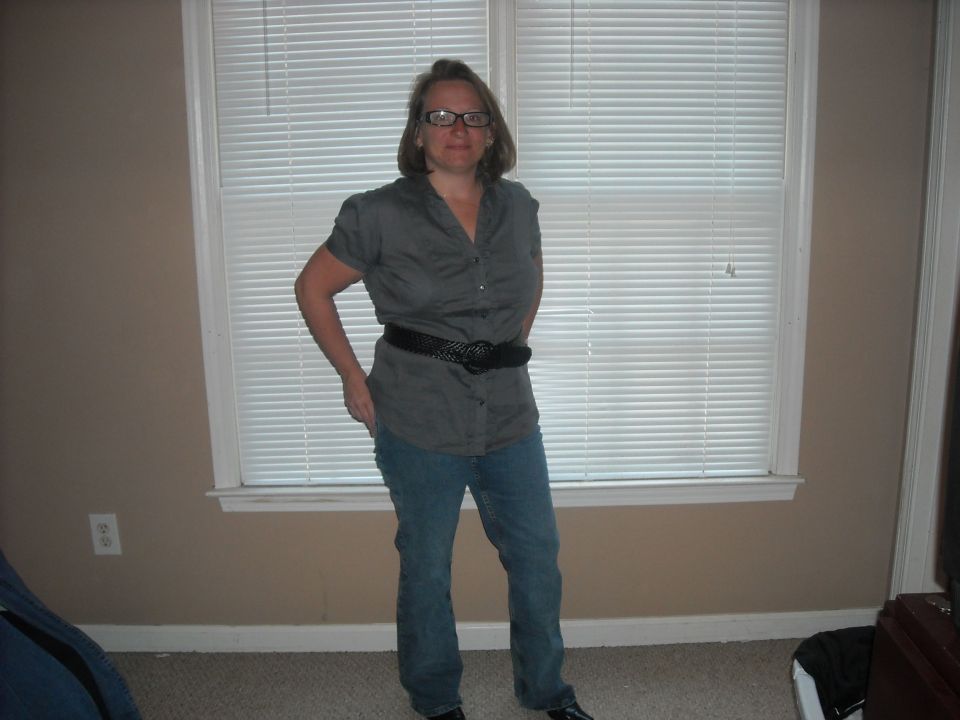 Woman with glasses and a grey shirt, black belt, and blue jeans stands in front of a window with blinds