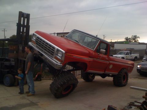 1989 Ford Bronco - A heavily modified red pickup truck is being lifted by a forklift, with two onlookers present.
