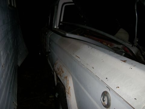 1976 Jeep J-10 - The side of a white pickup truck is visible, parked tightly between two structures. The hood is open, revealing the engine bay.