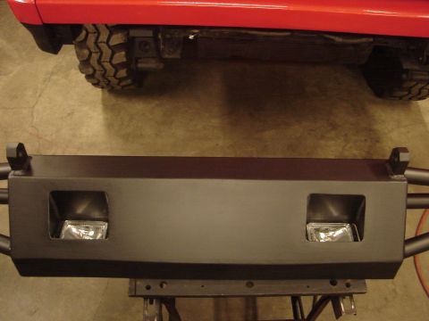 1989 Ford Bronco - A black aftermarket bumper with integrated fog light housings is shown, with a red vehicle's rear tire and fender visible above.