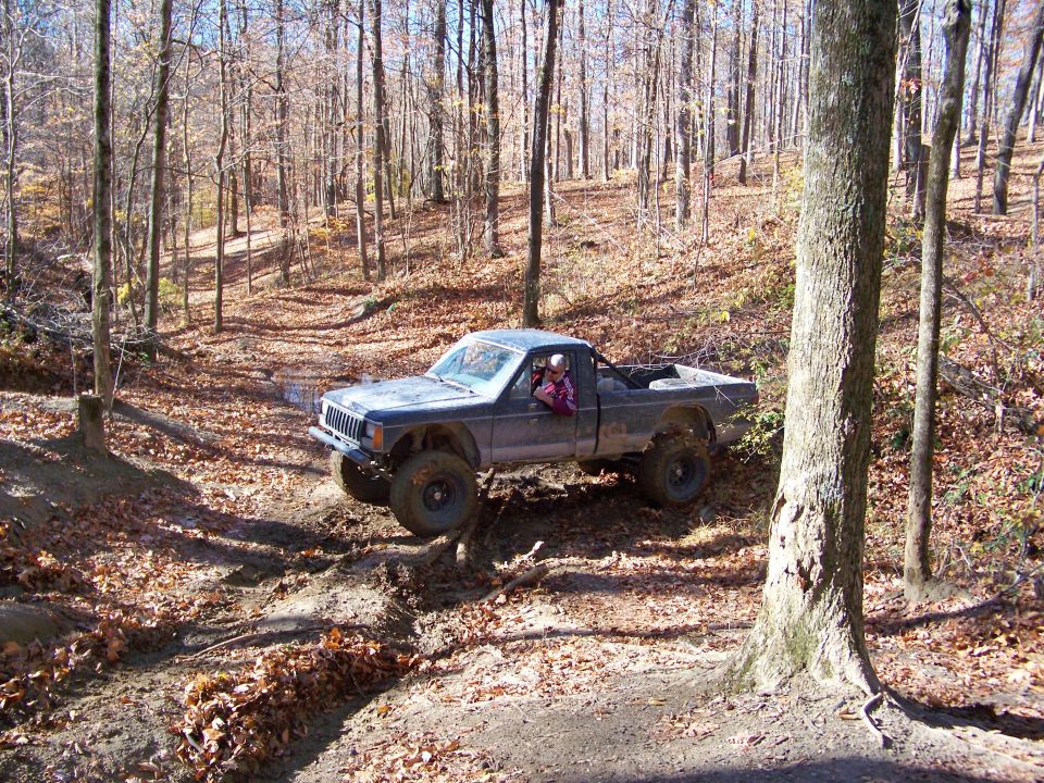 Jeep Comanche pickup truck with large tires driving on a dirt trail in a wooded area