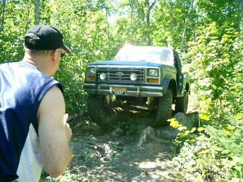 1986 Ford Bronco - A Ford Bronco tackles a challenging off-road course surrounded by dense forest foliage.