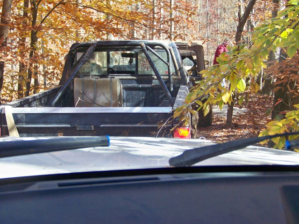 Pickup truck with roll cage and auxiliary fuel tank in wooded area. Another vehicle is partially visible behind it
