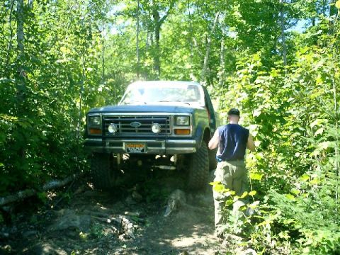1986 Ford Bronco - A Ford Bronco navigates a wooded trail, with a man observing its progress.