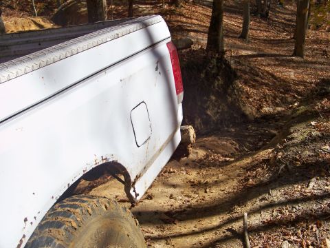 1987 Ford F-250 - A white pickup truck navigates a challenging, muddy off-road path surrounded by trees and fallen leaves.