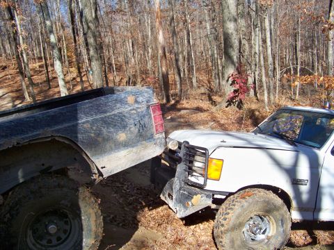 1987 Ford F-250 - Two pickup trucks are stuck bumper to bumper while off-roading in a wooded area.