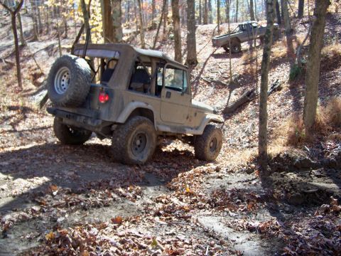 1987 Ford F-250 - A Jeep Wrangler tackles a challenging off-road path in a wooded area, with a second vehicle visible further up the incline.