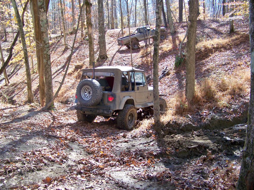 Tan Jeep Wrangler and a pickup truck driving on a wooded trail. The Jeep is in the foreground, facing away from the camera