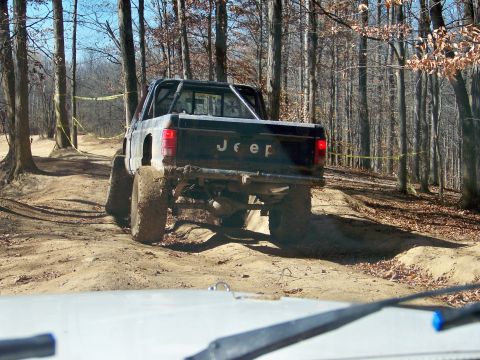 1987 Ford F-250 - A black Jeep pickup truck navigates a muddy trail through a wooded area.