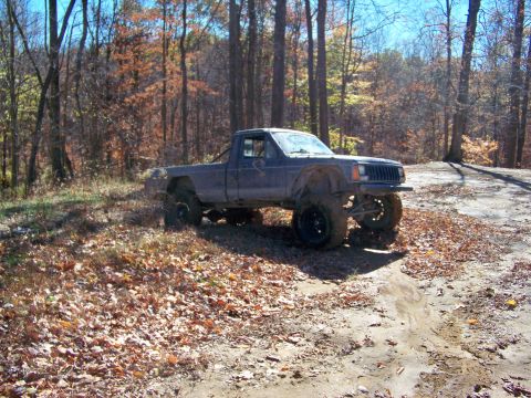 1987 Ford F-250 - A lifted Jeep Comanche pickup truck navigates a muddy trail surrounded by autumn trees.