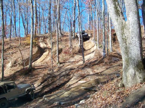 1987 Ford F-250 - Off-road vehicles tackle a challenging dirt path through a wooded area on a sunny day.