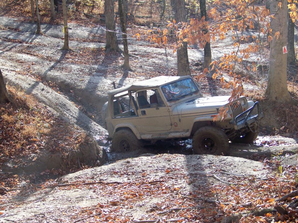 Tan Jeep Wrangler navigating a muddy trail in a wooded area