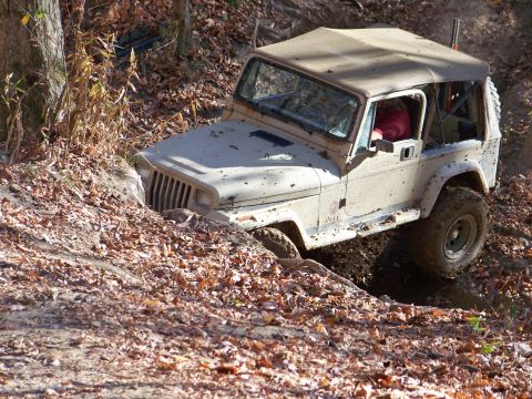 1987 Ford F-250 - A tan Jeep Wrangler navigates a challenging off-road trail, its tires deep in mud and surrounded by fallen leaves.