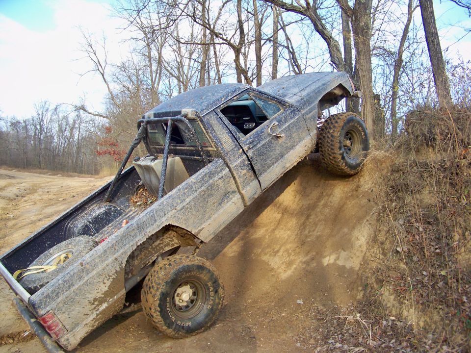 Mud-covered 1986 Jeep Comanche pickup truck with a roll cage, climbing a dirt hill