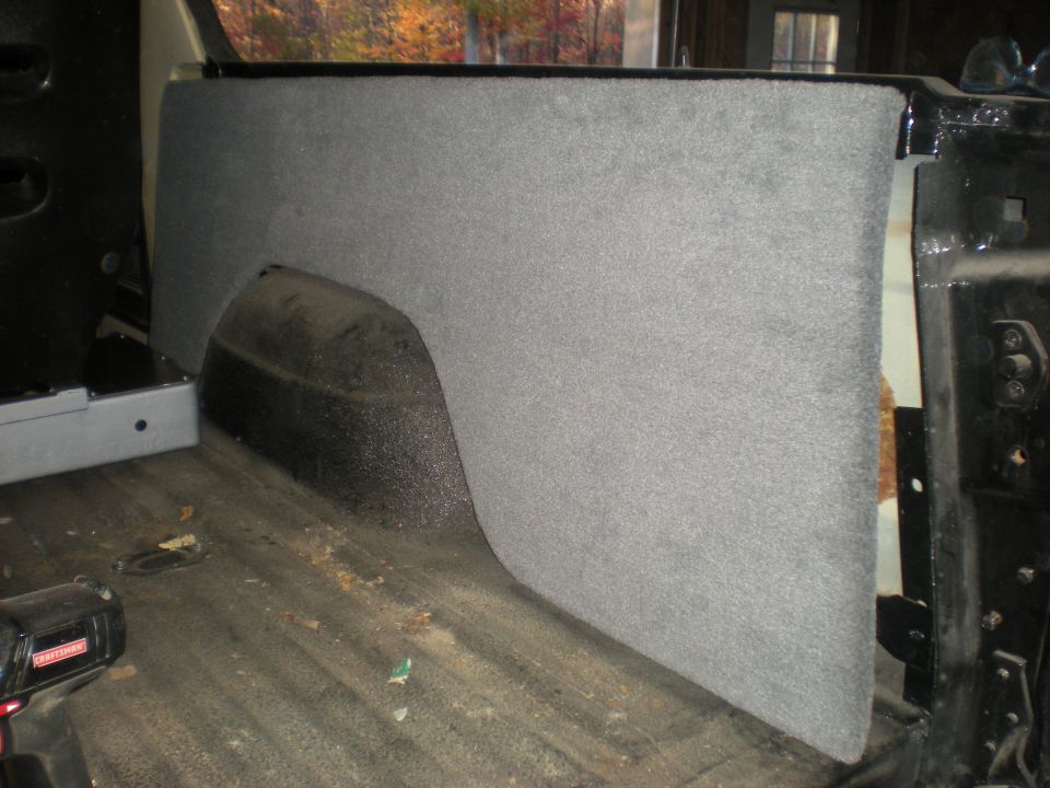 Interior rear quarter panel and wheel well covered in grey carpet. Floor shows dirt and a power tool