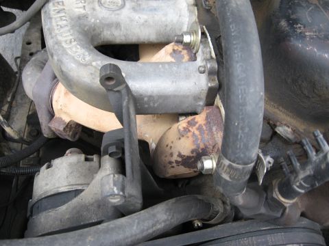 1981 Ford Bronco - Close-up of an engine bay showing cast aluminum intake or exhaust components with mounting bolts, along with rusty cast iron parts, black r…