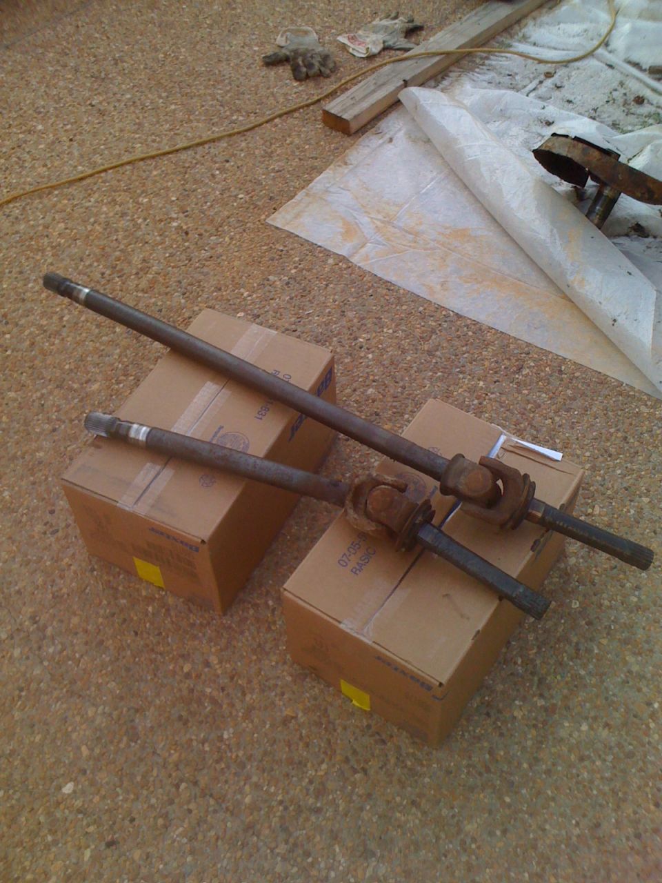Two drive shafts resting on cardboard boxes on a gravel surface. Some gloves and wood are in the background