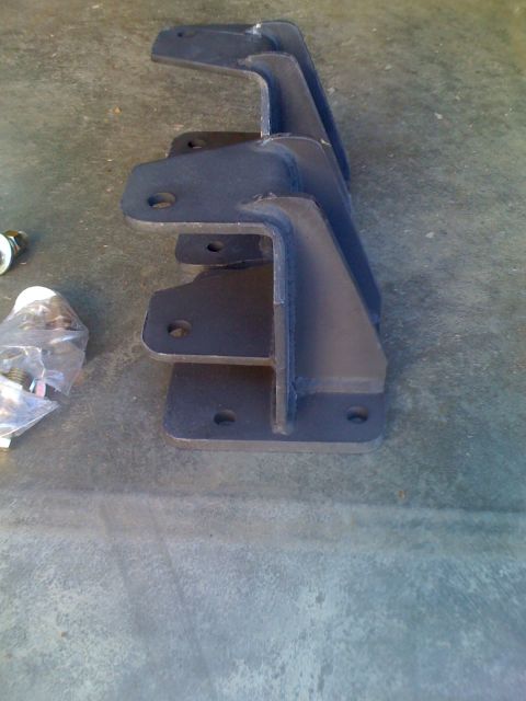 1992 Ford Bronco - Several dark metal suspension brackets are stacked on a concrete surface next to a bag of hardware.