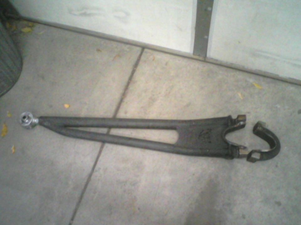 Dark gray metal automotive suspension control arm with a split end, lying on concrete