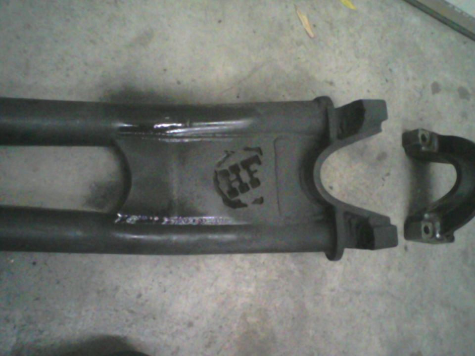 Close-up of a dark metal driveshaft component with a stamped logo that looks like 'HF'. Another bracket piece is to the right