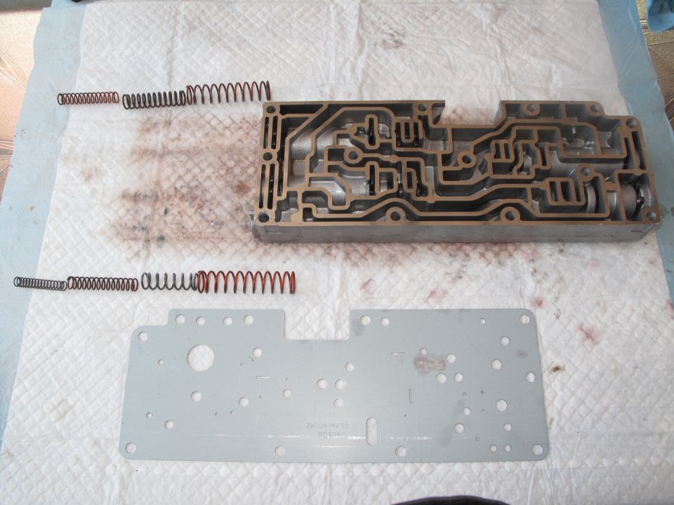 Transmission valve body and springs on a white cloth with red fluid stains. A metal plate is below the valve body