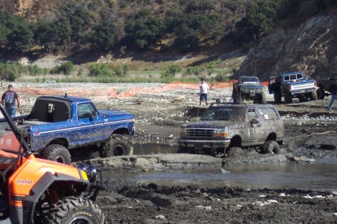 1995 Ford Bronco - Several off-road vehicles are stuck in a muddy terrain with trees and hills in the background.