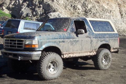 1995 Ford Bronco - Mud-splattered 1995 Ford Bronco parked off-road, with another vehicle visible to the left.
