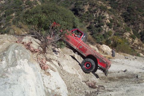 1995 Ford Bronco - A red off-road truck navigates a steep, rocky terrain with a person standing in the background.
