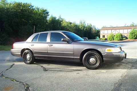 2001 Ford Crown Victoria - A gray 2001 Ford Crown Victoria sedan is parked on an asphalt surface with trees and a building in the background.