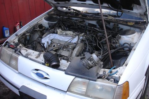 1990 Ford Taurus - The engine compartment of a white 1990 Ford Taurus is shown with the hood up, revealing various mechanical parts and wiring.