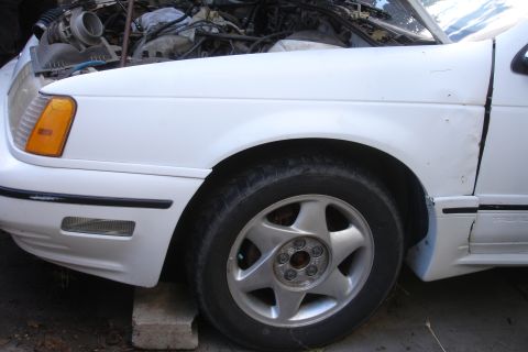 1990 Ford Taurus - The front fender and wheel of a white 1990 Ford Taurus are visible, with the hood open to reveal the engine compartment.