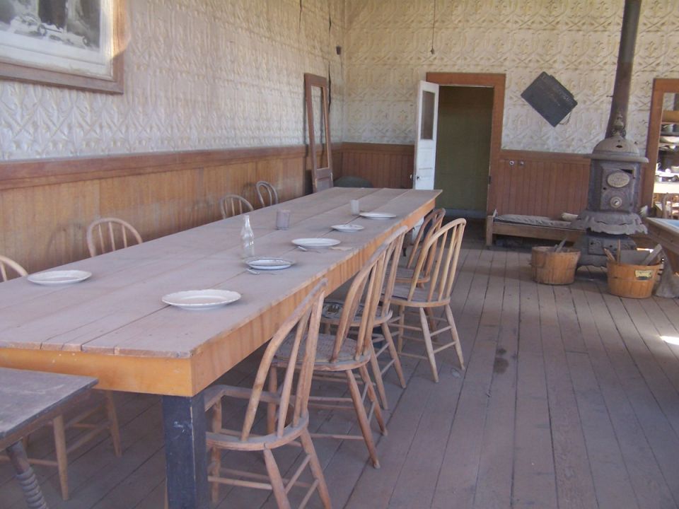 Long wooden table with chairs, set for a meal. Wood stove in corner. Old building interior