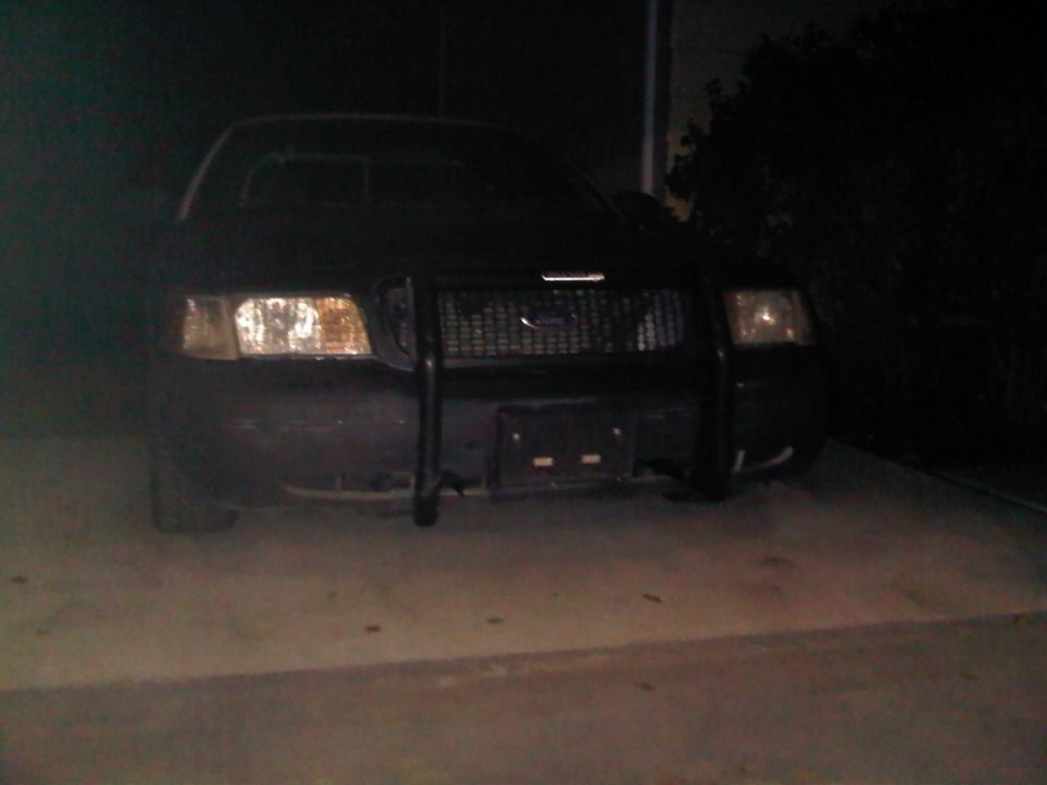 Front view of a 2003 Ford Crown Victoria Police Interceptor with a push bar. Headlights are on