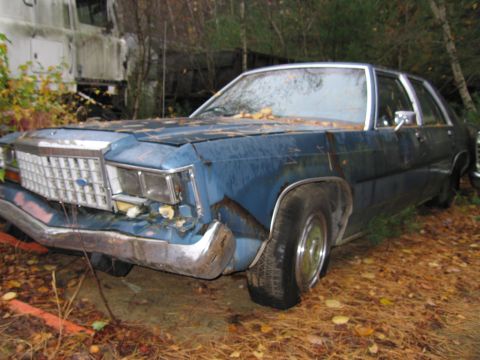 1991 Ford LTD Crown Victoria - A blue sedan sits in a junkyard, showing significant front-end damage and rust.