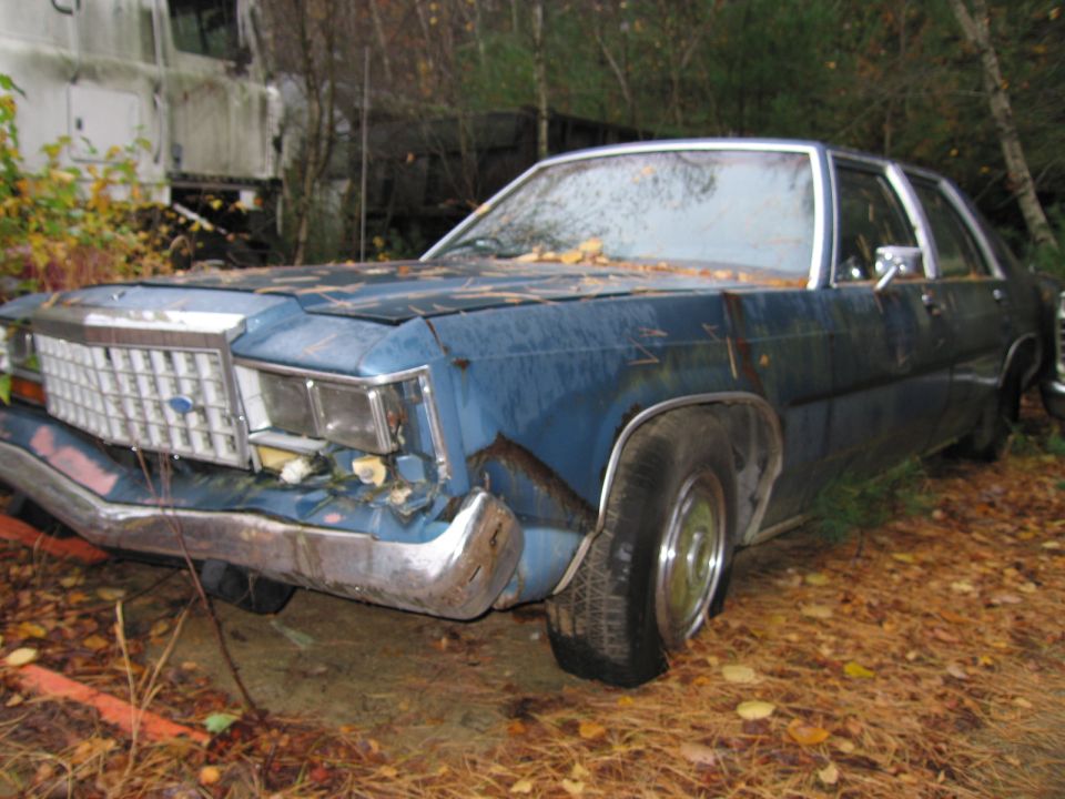 Blue sedan with dented front bumper and grille, parked in a junkyard