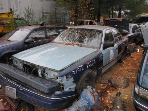 1991 Ford LTD Crown Victoria - This abandoned police car, likely a 1990s model, sits in a junkyard with visible damage and missing parts.