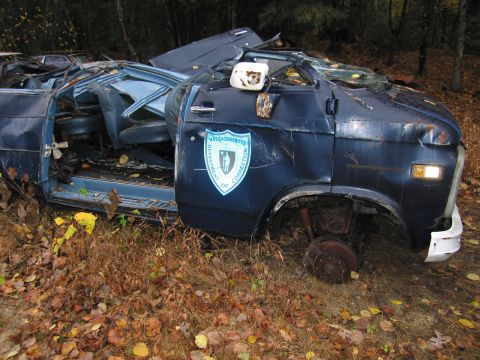 1991 Ford LTD Crown Victoria - This heavily damaged van, likely a former correctional vehicle, sits abandoned in a wooded area.