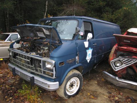 1991 Ford LTD Crown Victoria - A blue panel van sits in a junkyard with its hood open, next to another vehicle.