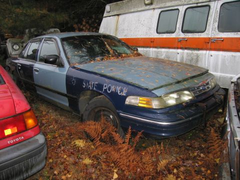 1991 Ford LTD Crown Victoria - This police car, possibly a 1991 Ford Crown Victoria LTD, is parked in a junkyard with other vehicles.