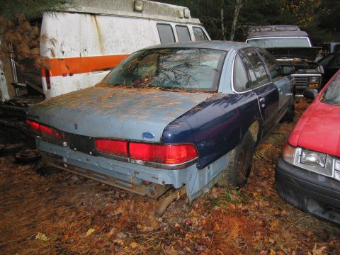 1991 Ford LTD Crown Victoria - A two-tone sedan sits in a junkyard next to an ambulance and other vehicles.