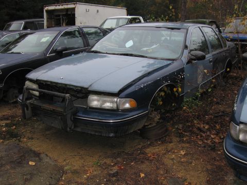 1991 Ford LTD Crown Victoria - A derelict blue sedan, missing its wheels and hood, sits in a junkyard.