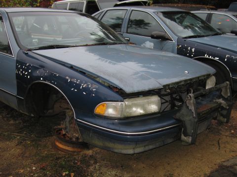 1991 Ford LTD Crown Victoria - A blue sedan sits in a junkyard, missing its wheels and front bumper, with other vehicles visible in the background.