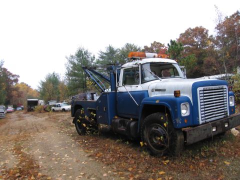 1991 Ford LTD Crown Victoria - An International tow truck sits in a junkyard, surrounded by fallen leaves and other vehicles.