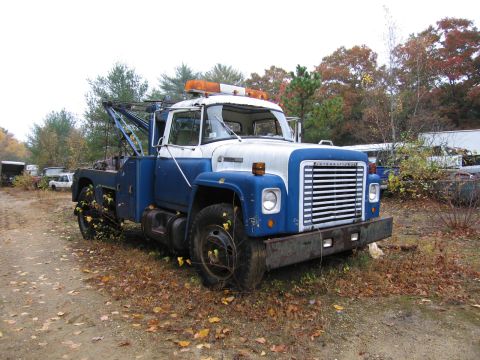 1991 Ford LTD Crown Victoria - An old International Harvester tow truck sits in a junkyard with fallen leaves on the ground.