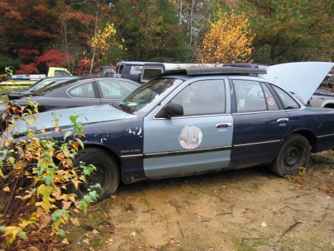 1991 Ford LTD Crown Victoria - Side view of a 1991 Ford Crown Victoria LTD police car in a junkyard with other vehicles visible.