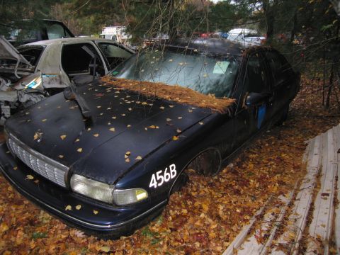 1991 Ford LTD Crown Victoria - A dark sedan sits in a junkyard, covered in leaves and missing a front wheel.