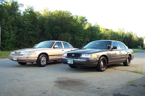 2001 Ford Crown Victoria - Two Ford Crown Victorias, a tan one and a gray one, are parked outdoors on an asphalt surface with trees in the background.