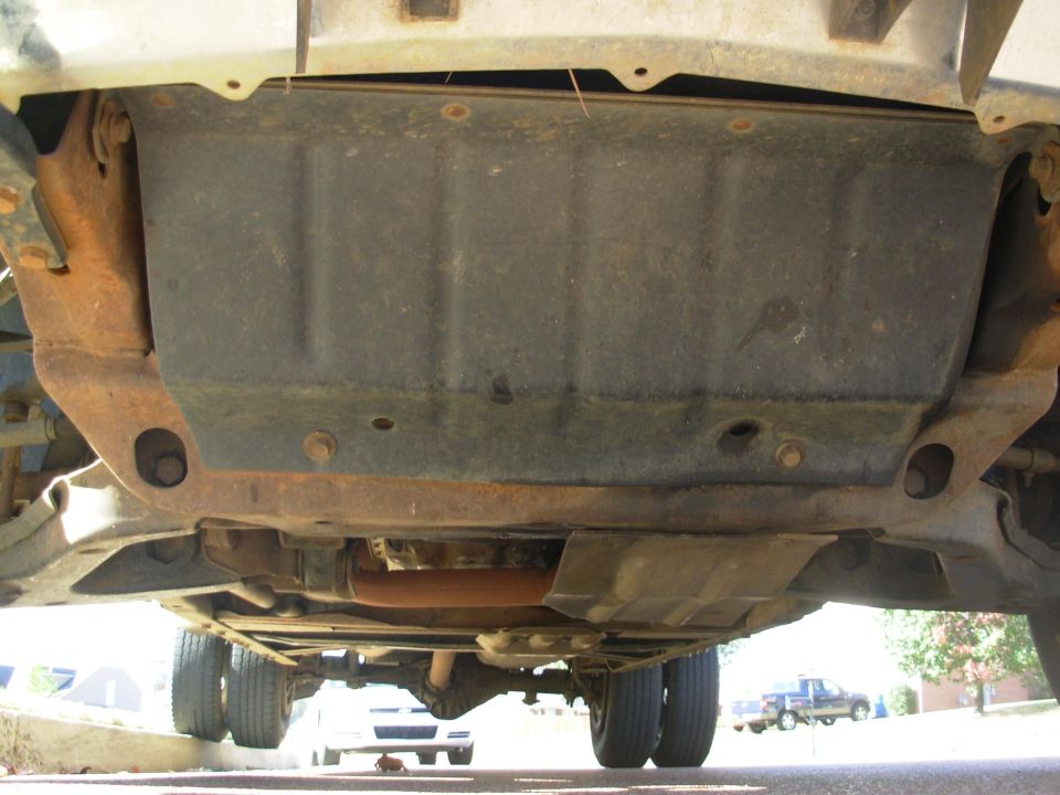Under-truck view of a black skid plate, rusty crossmember, exhaust, and dual rear wheels on pavement
