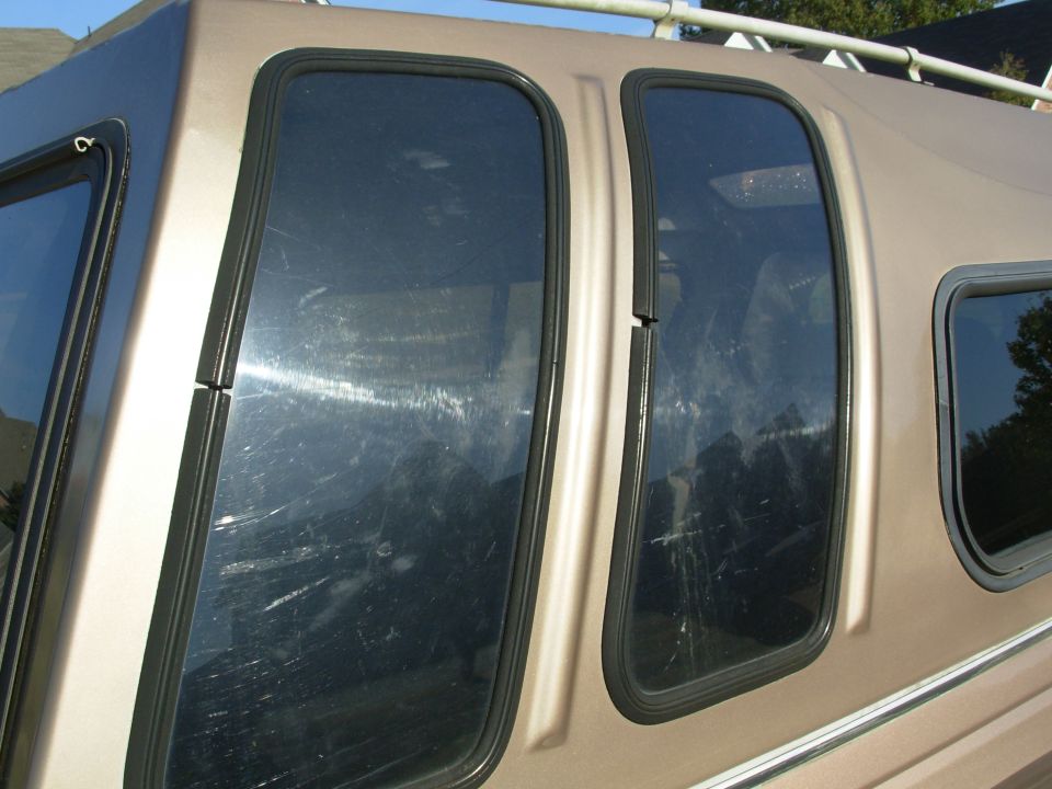 Gold camper shell side windows with black trim and roof rack, tinted glass reflecting trees