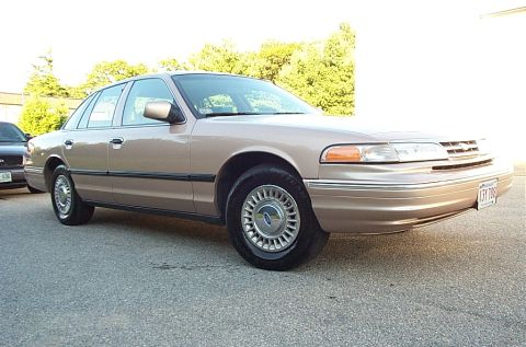 2001 Ford Crown Victoria - A 2001 Ford Crown Victoria is parked on asphalt, with trees and a building in the background.